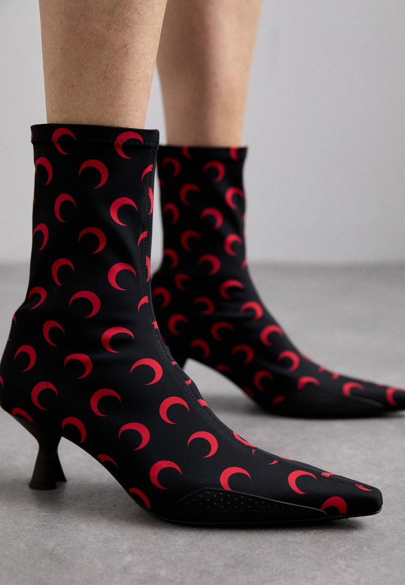 Botines negros de tobillo hechos de material elástico, con punta afilada y tacón kitten. Decorados con patrones de luna creciente roja en todo su diseño.