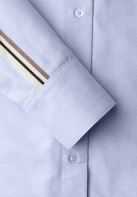 Lyseblåt skjortestof med en glat tekstur. Manchetten har et stribet design i brun, creme og sort. Knaplukning ved manchetten.