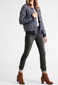 Marineblaue gemusterte Bomberjacke mit rundem Strickkragen, kombiniert mit grauen Skinny-Jeans und braunen Wildleder-Ankle-Boots mit Blockabsatz.