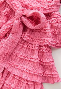 Rózsaszín, fodros textil, amelynek textúrált felülete van. Tartalmaz egy megkötött masni részletet és rétegezett fodrokat, amelyek egyenletes mintát mutatnak az egész anyagon.