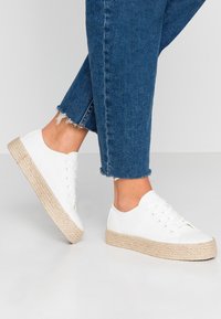 Sneakers bianchi in tela con suola in juta intrecciata naturale, dotati di lace-up e punta arrotondata. Indossati con jeans in denim blu sfilacciati.