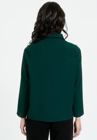 Zaļa, viegla blūze ar apkakli, garām piedurknēm un noapaļotu apakšmalu. Gluda tekstūra bez redzamām rakstiem vai akcentiem.