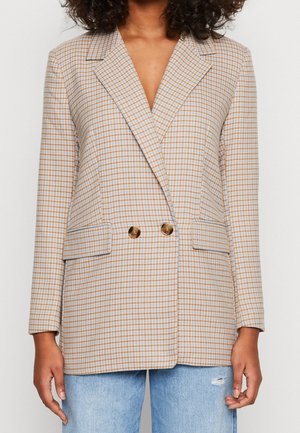 Kobieta ubrana w beżowo-brązową dwurzędową marynarkę w kratę z złotymi guzikami oraz jasnoniebieskie podarte dżinsy.