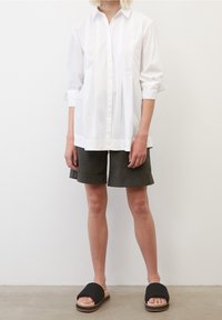 Camisa blanca de manga larga con tapeta de botones, combinada con pantalones cortos oscuros verdes y sandalias de slide negras con suela de madera.
