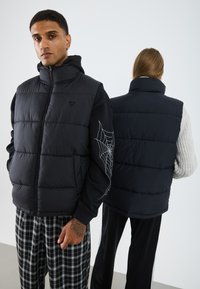 Gilet matelassé noir avec col montant, fermeture éclair à l'avant et design matelassé. Présente un motif de toile d'araignée sur la manche gauche. Le dos est lisse et uni.
