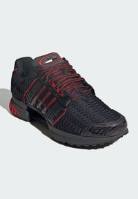 Black athletic shoe with red accents, textured knit fabric, plastic overlays, a padded collar, and a rubber outsole featuring grip patterns.