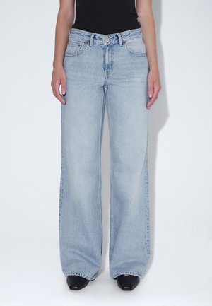 Osoba ubrana w jasnoniebieskie szerokie jeansy, czarne spiczaste buty oraz czarny top bez rękawów na białym tle.