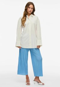 Cămașă albă cu butoni, cu mâneci lungi, asortată cu pantaloni largi plisati de un albastru deschis și sandale cu toc albe. Design simplu și modern.