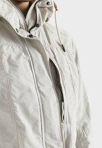 Chaqueta de color beige claro con cuello alto, cremallera frontal y bolsillo lateral. El material tiene una textura suave con detalles de costura visibles.