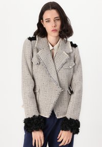 Lichtgrijze tweed blazer met rafelige randen, dubbele knoopsluiting, subtiele glans en zwarte bloemenaccenten op de mouwen.