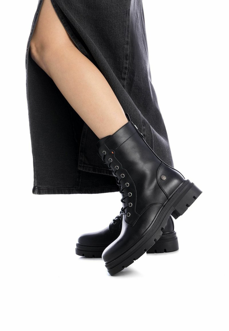 Botas de combate de cuero negro con cordones, suela gruesa y herrajes metálicos, que presentan una textura lisa y un diseño de punta redonda.