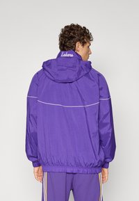 Lila szélfogó dzseki kapucnival, rugalmas mandzsettákkal és derékpánttal, fehér szegéllyel és a nyakán hímzett "Lakers" felirattal.
