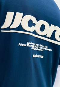 Camiseta de algodón azul con gran nombre de marca y texto impresos en blanco. El texto está situado en la parte trasera. Textura suave, diseño casual.