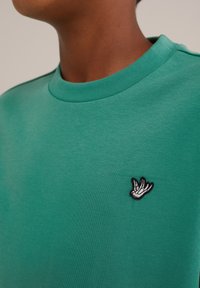 Žalias medvilninis marškinėliai su apvalia iškirpte ir mažuoju juodu bei baltu siuvinėtu paukščio logotipu krūtinėje. Lygi tekstūra.