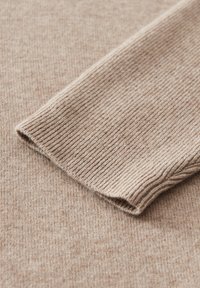 Beiger gerippter Strickstoff mit umgeschlagenem Ärmelsaum, der die weiche Textur und die feine Naht des Materials hervorhebt.