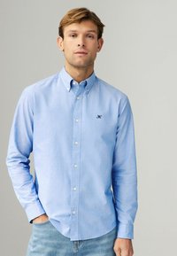 Camisa de botones azul claro en mezcla de algodón, de mangas largas, con un pequeño logo bordado en el pecho. Textura suave, diseño clásico de cuello.