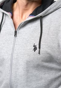 Harmaa vetoketjullinen huppari, jossa on musta sisähuppu ja punasinivalkoinen viimeistely, ja rinnassa pieni musta kirjailtu logo.