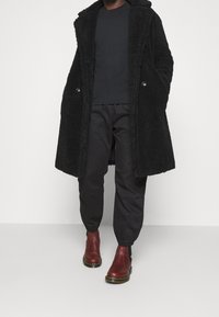 Svart fleecejacka med huva, två framfickor och knäppning, som matchas med svarta byxor med elastiska manschetter och burgundy ankelskor.