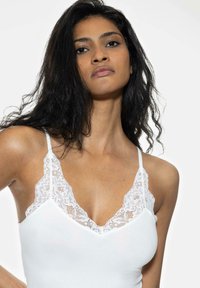 Valkoinen toppi, jossa on pitsikoristelu v-pääntiellä ja olkaimissa, ominaisuuksina pehmeä, sileä rakenne ja tiukka muotoilu.