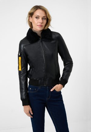 Giacca bomber in pelle nera con colletto morbido nero, tasca con zip e accentuazione gialla, polsini e vita a costine, abbinata a jeans blu.