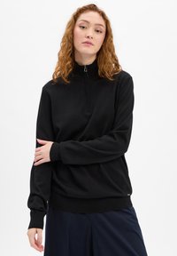 Schwarzer Reißverschluss-Pullover mit hohem Kragen, langen Ärmeln, gerippten Bündchen und Saum, hergestellt aus einem weichen Stoff. Bemerkenswert durch sein minimalistisches Design.