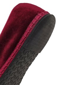 Acercamiento de una pantufla de terciopelo burdeos con bordes cosidos y una suela de goma texturizada negra.