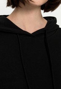 Sweatshirt à capuche noire en tissu doux, avec un décolleté profond et des coutures visibles sur la capuche et le corps, créant un design décontracté.