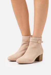 Bottines en daim beige avec bout pointu, une fermeture éclair latérale et une lanière décorative à boucle. Elles possèdent un talon en bois empilé.