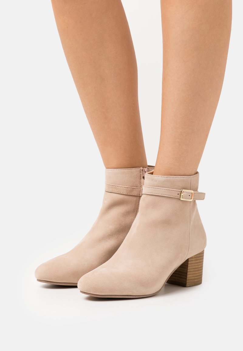 Bottines en daim beige avec bout pointu, une fermeture éclair latérale et une lanière décorative à boucle. Elles possèdent un talon en bois empilé.