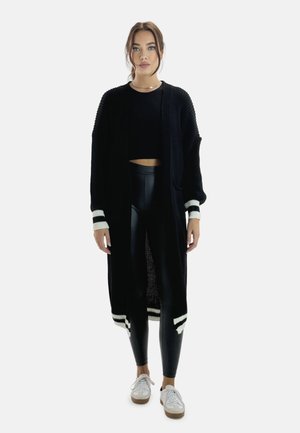 Sort strik cardigan med hvide stribede manchetter, båret over en sort cropped top og skinnende sorte leggings, parret med hvide sneakers.