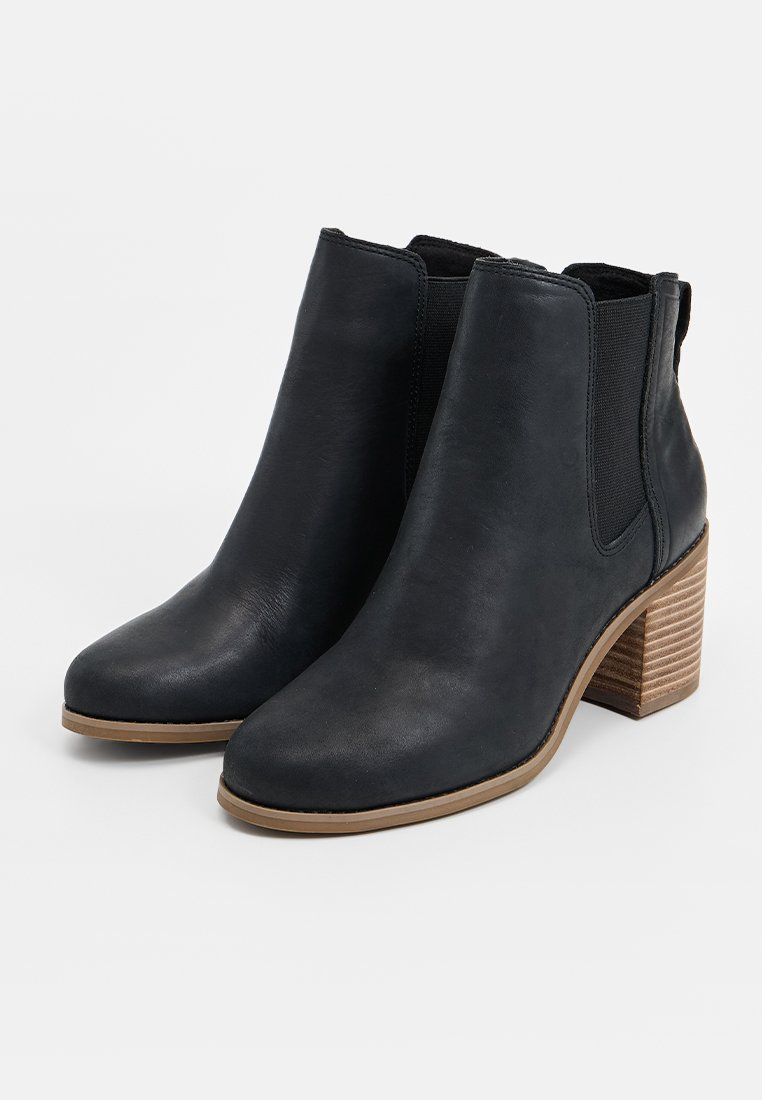 Zwarte leren enkellaarzen met een blokhak en elastische zijpanelen, met een gladde textuur en ronde neus, geplaatst op een lichtbruine zool.