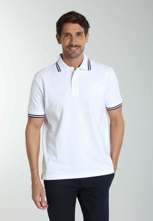 Homme souriant portant un polo blanc avec des rayures noires sur le col et les manches, et un pantalon foncé, debout devant un fond clair.