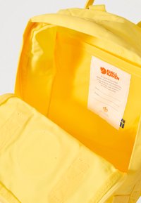 Dzeltena mugursoma no izturīga neilona materiāla, ar plašu galveno nodalījumu, kurā ir etiķete un gluda tekstūra; aprīkota ar diviem priekšējiem rāvējslēdzējiem.