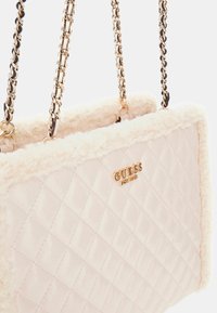 Borsa a tracolla trapuntata di colore rosa chiaro con rifiniture in pelliccia sintetica crema, catene dorate e logo "GUESS" in oro sulla parte anteriore. Texture morbida e design strutturato.