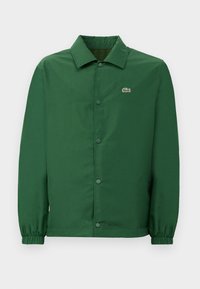 Chaqueta verde con un cuello, botones de presión en el frente y puños elásticos. Presenta un pequeño logo bordado en el lado izquierdo del pecho. Textura suave.