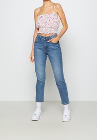 Kvinna som bär en blommig ärmlös kort topp, blå högmidjade jeans, vita strumpor och vita sneakers, stående med ena handen i fickan.