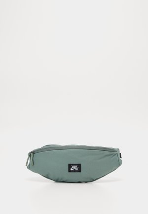 Sac banane vert Nike SB avec fermeture éclair et patch logo rectangulaire noir sur le devant, présenté sur fond blanc uni.