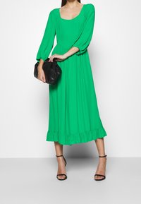 Vihreä midi-mekko, jossa on pitkät puffihiat, röyhelöhelma ja tiukka vyötärö. Yhdistetty mustan laukun ja mustien soljillisten korkokenkien kanssa.