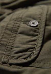 Tejido verde olivo con un acabado texturizado, que presenta un bolsillo de solapa y un detalle de botón de metal, resaltado por costuras en contraste.