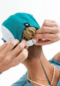 A teal and white cap made of mesh material, adjustable at the back with a white plastic strap and a black label on the side.