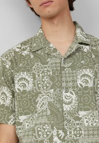 Camicia a maniche corte con bottoni di colore verde oliva, decorata con intricati motivi floreali e geometrici bianchi. Tessuto leggero con una vestibilità rilassata.