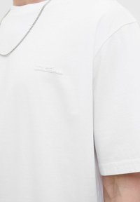Hvid bomulds t-shirt med korte ærmer, prydet med et tekstureret logo-patch og en rund halsudskæring. Akkompagneret af et sølv-kæde halskæde.