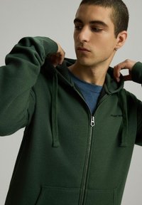 Zielona bluza zapinana na zamek z kapturem na sznurku, o gładkiej fakturze i przednich kieszeniach. Założona na niebieską koszulkę z okrągłym dekoltem.