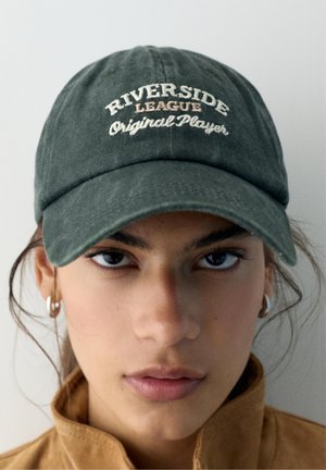 Nuori henkilö ja korvarenkaat, joka käyttää vihreää lippalakki jossa kirjailtuna "Riverside League Original Player" ja ruskeaa takkia, jonka kaulus on ylös käännetty.