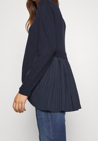 Námořnicky modrý svetr s dlouhými rukávy, zdobený plisovaným vzorem na zádech a žebrovanými manžetami, doplněný modrými džíny. Celkově hladká textura.