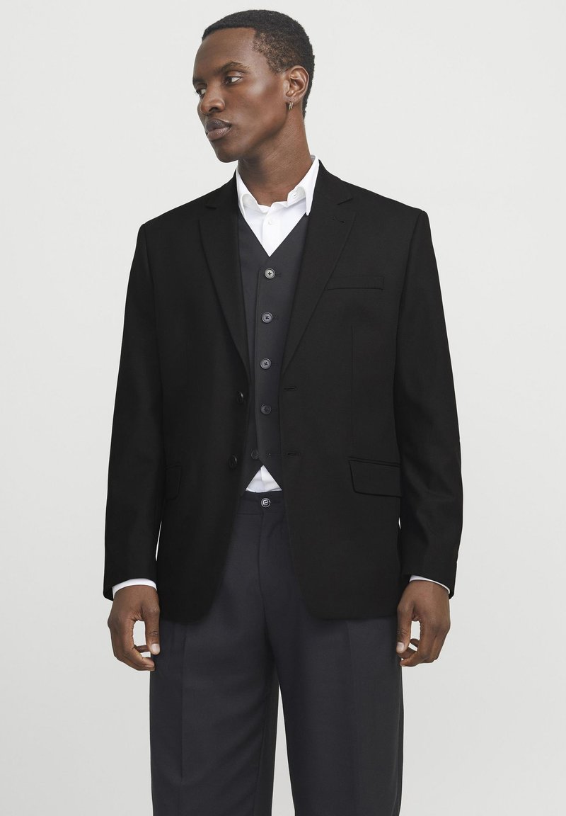 Zwarte op maat gemaakte blazer met een notch-revers, een enkelvoudige knoopsluiting aan de voorkant en twee klepzakken, gedragen over een grijs vest en een wit shirt.