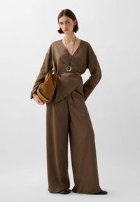 Bruine wikkeltop met een v-hals en lange mouwen, gecombineerd met brede bruine pantalons. Het model houdt een bruine tas vast, vervaardigd uit een gestructureerde stof.