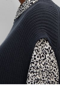 Haut sans manches en tricot gris foncé superposé à une chemise à motifs noir et blanc avec col. Tissu texturé et coupe ample.