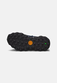 Suela de goma negra con surcos profundos, logo naranja en el centro y acento verde. Diseñada para tracción y durabilidad.