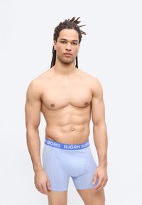 Jeune homme avec des dreadlocks portant un boxer Björn Borg bleu clair, debout devant un fond gris clair uni.